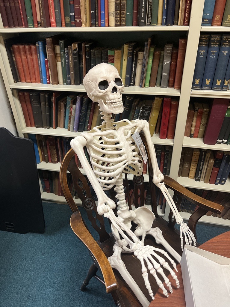A skeleton sits in a chair. They are surrounded by old books. 